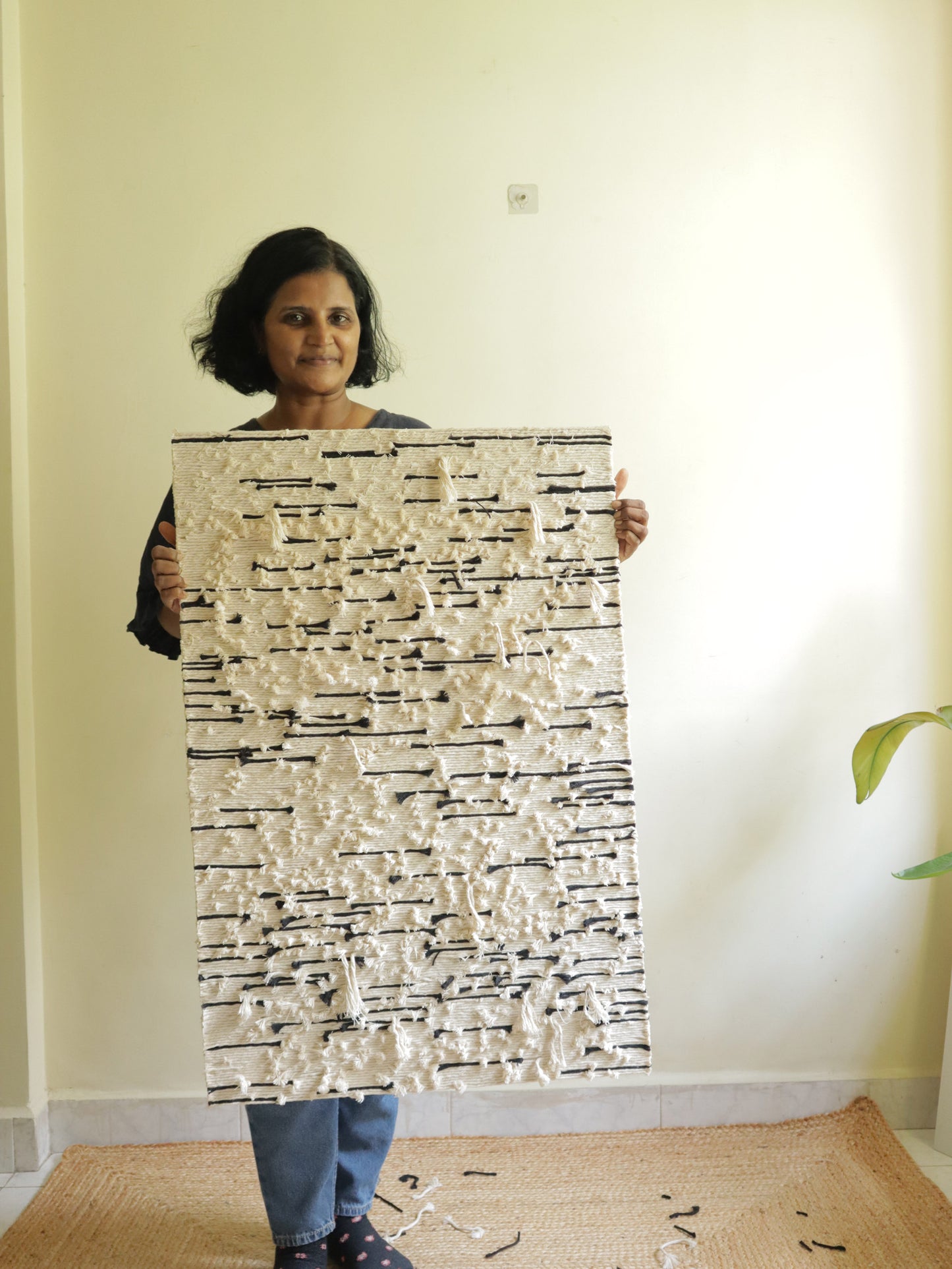 Person holding a large abstract artwork in a room with a light-colored wall and a plant.