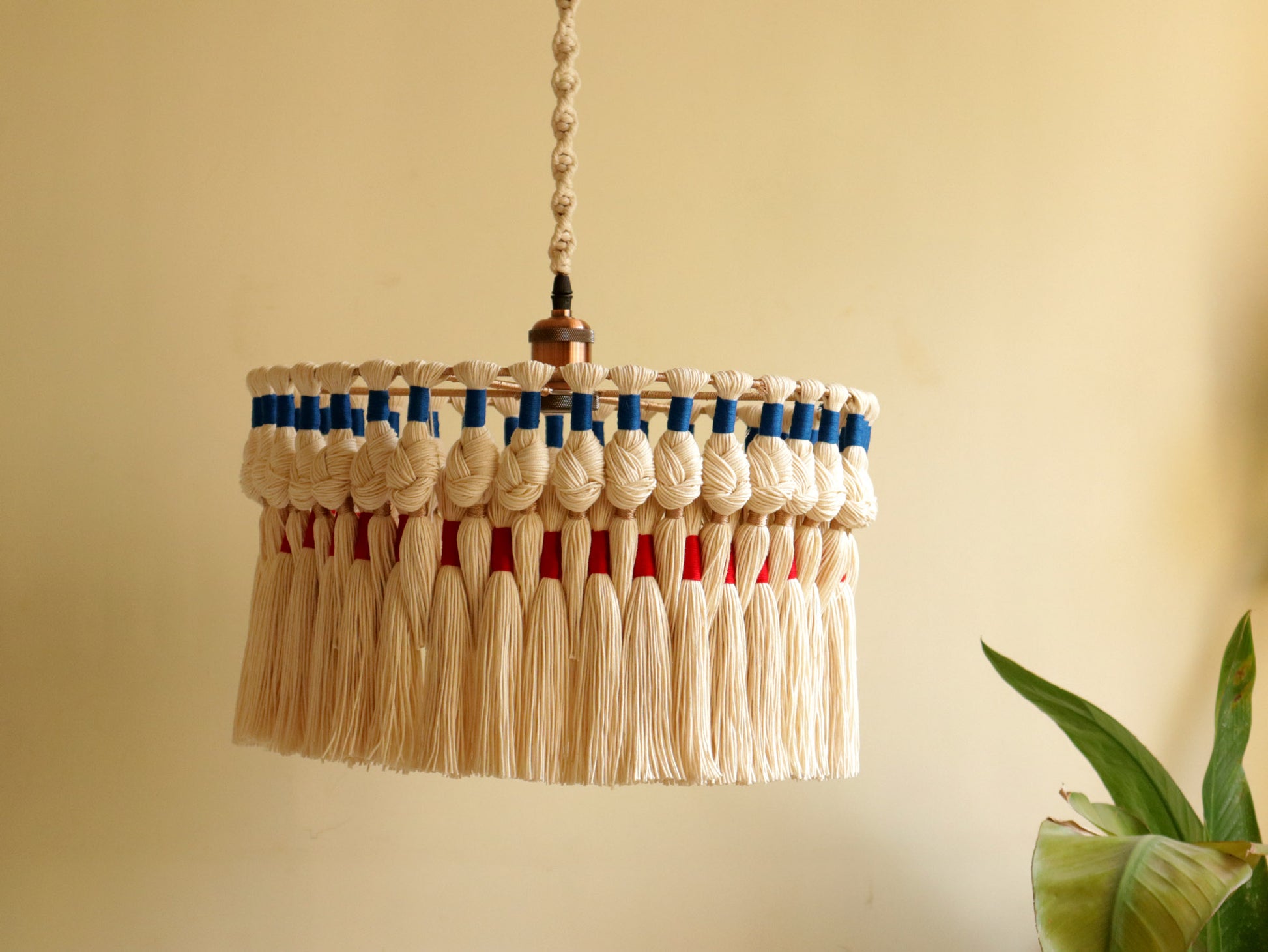 May include: Close-up of a cream-colored macrame lampshade with blue and red accents. The lampshade features knotted details and long, fringed tassels. The design is handmade and adds a bohemian touch to home decor.